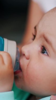 Güzel beyaz çocuk şişeden su içiyor ve gülümsüyor. Annenin eli oğlu için bir şişe tutuyor. Kapatın. Dikey video