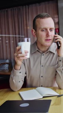 Erkek ofis çalışanı telefonda konuşuyor ve çay içiyor. Genç adam güzel modern ofisin arka planında masada oturuyor. Dikey video