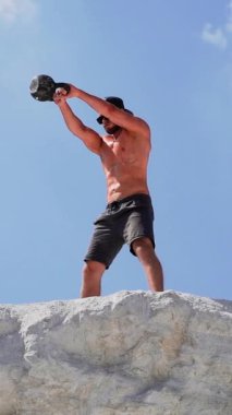 Dışarıda kettlebell sallayan sağlıklı bir sporcu. Spor salonundaki adam, beyaz dağın tepesinde antrenman yaparken ağır kettlebell ağırlığını kaldırıyor. Dikey video