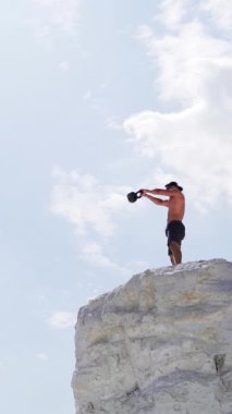 Tepede ağır bir yük taşıyan sağlıklı bir adamın eğitimine bir de aşağıdan bak. Vücut geliştirici, doğal gökyüzü arka planında kettlebell kaldırıyor. Spor yaparken gömleksiz bir adam. Dikey video