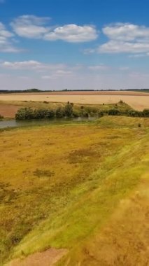 Çayırları tarlalardan ayıran uzun bir tepe. Tarlaların ortasında küçük bir gölet. Arka planda bulutlar olan mavi gökyüzü. Dikey video