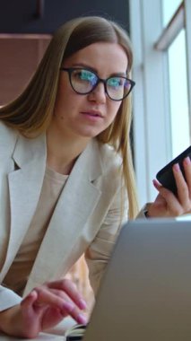 Sarışın genç bayan telefonda konuşuyor. Masaya yaslanıyor. Kadın çalışan telefonda konuşur ve dizüstü bilgisayarla çalışır. Ofis arka planda. Dikey video