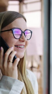 Telefonda konuşan gülümseyen bayan. Büyük panoramik pencerenin yanında dikey duran genç bir bayanın portresi.