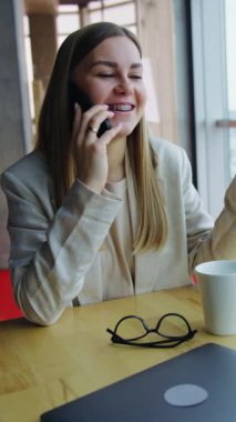 Genç bayan telefonda hoş sohbetler yapıyor ve gülümsüyor. Öğle yemeğinde çay içip, akıllı telefondan konuşmak. Şehir manzarası arka planda. Dikey video