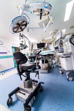 Geniş bir tepe lambası ve tıbbi teçhizatı olan bir hastane odası. Oda temiz ve aydınlık, profesyonellik ve bakım hissi veriyor.