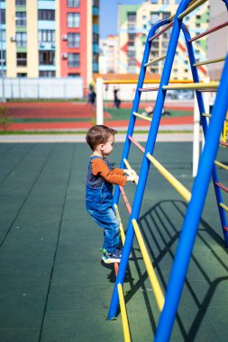 Genç bir çocuk mavi ve sarı bir merdivene tırmanıyor. Sahne arka planında renkli bir bina olan bir parkta geçiyor.