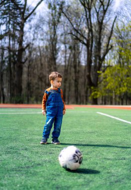 Mavi tulumlu genç bir çocuk elinde beyaz bir futbol topuyla bir futbol sahasında duruyor. Çocuk topa bakıyor, muhtemelen tekmelemeye hazırlanıyor.