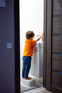 Genç bir çocuk banyoda dişlerini fırçalıyor. Çocuk turuncu bir gömlek ve kot pantolon giyiyor.