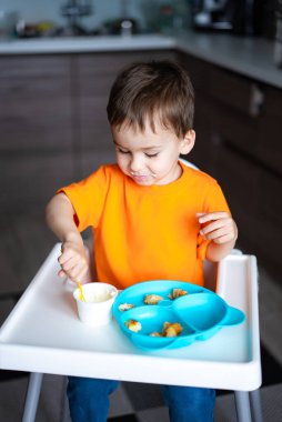 Genç bir çocuk yüksek bir sandalyede oturup yemek yiyor. Turuncu bir gömlek giyiyor.