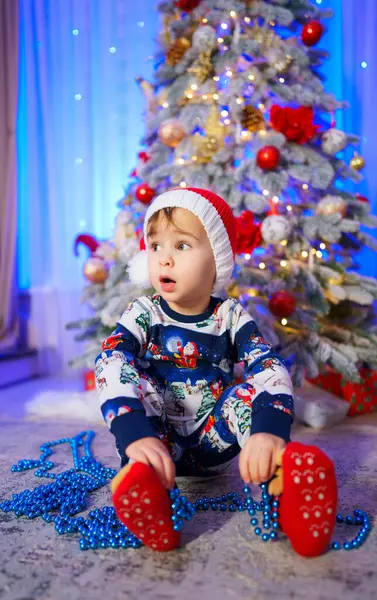 Lille Barn Sidder Foran Juletræ Iført Julemandshat Blåt Hvidt Tøj Royaltyfrie stock-fotos