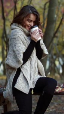 Parkta kahve içen bir kız. Sonbahar ormanında elinde bir fincan kahve tutan genç bir kadın. Dikey video.