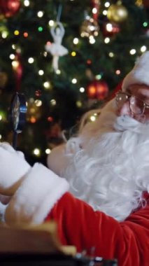 Noel Baba büyüteçten zarfa bakıyor. Noel ağacı arkaplanı ve beyaz sakallı Noel Baba zarftaki adresi okuyor. Noel zamanı. Dikey video