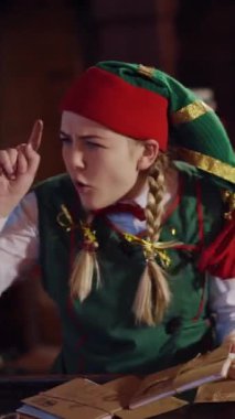Kutlama arka planında zarfları olan komik bir elf. Yeşil ve kırmızı kostümlü neşeli kadın elf Noel Baba için zarflara bakıyor ve mutlu bir şekilde dans ediyor. Dikey video