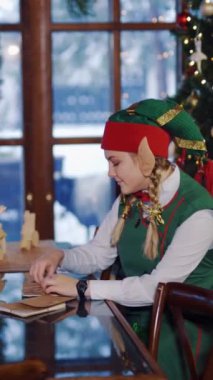 Kutlama masasında zarfları olan bir elf. Noel ağacı arka planında neşeli bir elfin zarf açtığı yan görüntü. Dikey video
