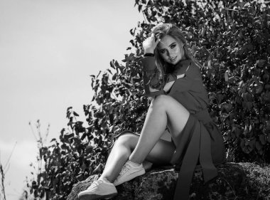 Bacak bacak üstüne atmış, elinde saçı olan bir kadın bir kayanın üzerinde oturuyor. O kırmızı bir ceket ve beyaz ayakkabılar giyiyor..