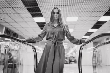 Uzun elbiseli bir kadın yürüyen merdivenin önünde duruyor. Yürüyen merdiven boş ve sahnedeki tek kişi kadın.