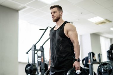 Siyah kolsuz tişörtlü bir adam elinde iki dambıl tutuyor. Spor salonunda çalışırken odaklanmış ve kararlı görünüyor.