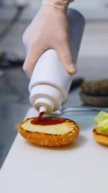 Cook adding ketchup on burger. Preparing burger on kitchen. Cook hands in white gloves preparing and making hamburger. Selective focus. Vertical video