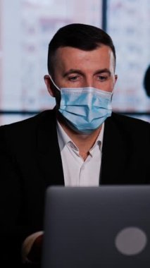 Cerrahi maske takan bir iş adamı dizüstü bilgisayarda çalışıyor. Büyük bir ofiste bilgisayarın başında oturan bir adam. Arkaplan bulanık. Dikey video