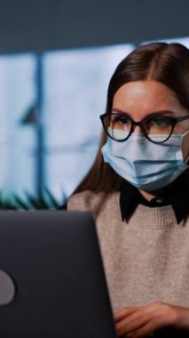 Maskeli kadın ofiste dizüstü bilgisayarında çalışıyor. İş kadını bilgisayarı kapatıyor, gözlüklerini çıkarıyor ve maskesini takıp bilgisayara takıyor. Dikey video