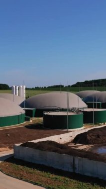 Modern biyogaz üretim tesisi. Enerji üretimi için yuvarlak depolama tankları. Arkaplanda yeşil tarlalar, ormanlar ve mavi gökyüzü. Dikey video