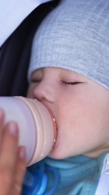 Gri şapkalı ve ceketli güzel çocuk şişeden süt içiyor. Sağlıklı erkek bebek uykuya dalmak için gözlerini kapattı. Kapatın. Dikey video