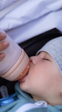 Bebek arabasında yürürken tapılası bebeği beslemek. Tatlı çocuk süt emiyor ve yavaş yavaş uykuya dalıyor. Kapatın. Dikey video