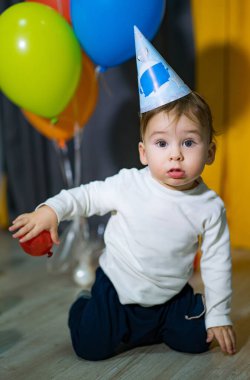 Zürafa tişörtü ve parti şapkası giymiş bir bebek yerde oturuyor. Bebek kırmızı bir balon tutuyor.