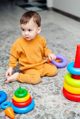 Genç bir çocuk, bir yığın blok ve bir dizi yüzük de dahil olmak üzere çeşitli oyuncaklarla çevrili olarak yerde oturuyor..