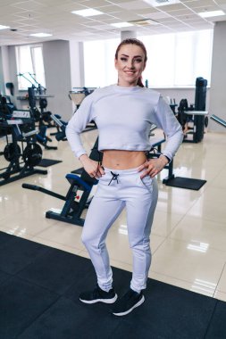 Beyaz gömlekli ve beyaz pantolonlu bir kadın spor salonunda poz veriyor. Gülümsüyor ve mutlu.