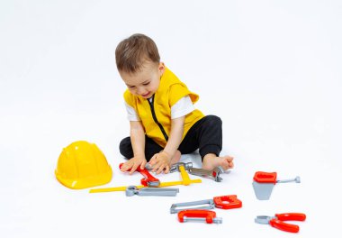 Küçük bir çocuk çekiç ve ingiliz anahtarı da dahil olmak üzere bir dizi oyuncak aletle oynuyor. Çocuk sarı bir yelek ve sarı bir kask giyiyor..