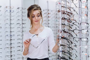 Bir kadın mağazada bir çift gözlüğe bakıyor. Elinde bir çift gözlük tutuyor ve onları deniyor gibi görünüyor..
