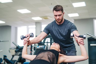 Bir erkek bir kadına antrenmanında yardım ediyor. Adam bir çift dambıl tutuyor ve kadın bir bankta yatıyor.