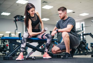 Bir erkek ve bir kadın spor salonundalar. Bir erkek, kadının antrenmanına yardım ediyor.