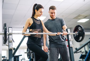 Bir adam ve bir kadın spor salonundalar, halter tutan bir adam ve ona bakan bir kadın..