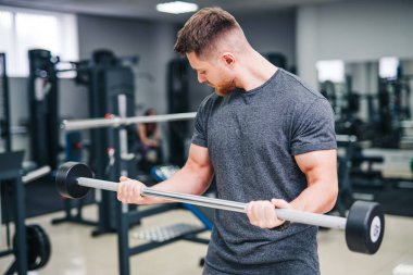 Bir adam spor salonunda ağırlık kaldırıyor. Gri bir gömlek giyiyor ve sakalı var.
