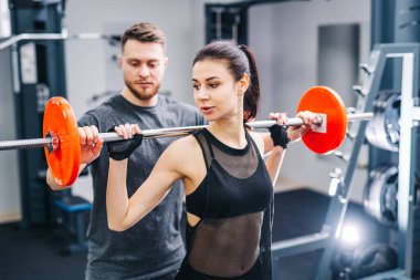 Bir erkek ve bir kadın spor salonunda ağırlık kaldırıyorlar. Adam kadına formunda yardım ediyor.