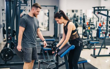 Bir erkek ve bir kadın spor salonundalar, erkek kadına makinenin nasıl kullanılacağını gösteriyor.
