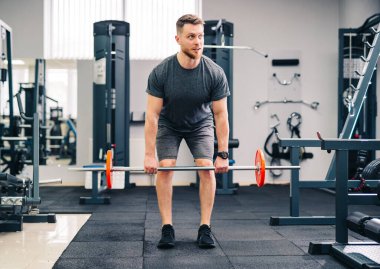 Bir adam spor salonunda halter kaldırıyor. Siyah bir gömlek ve gri bir şort giyiyor.