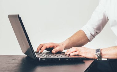 Bir adam dizüstü bilgisayarda yazıyor. Laptop siyah ve gümüşten. Adam beyaz bir gömlek giyiyor.