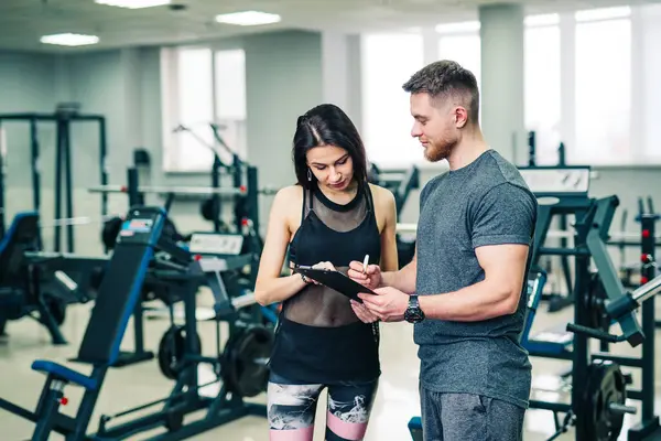 Bir erkek ve bir kadın spor salonundalar, erkek tablete bir şeyler yazarken kadın izliyor.