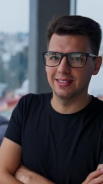 Gülümseyen koyu renk saçlı genç adam gözlüklerini çıkarıyor. İşadamı boş bir ofiste dikilip pencereden dikkatlice bakıyor. Dikey video