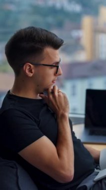 Ofiste oturup iş meselelerini düşünmek. Fasulye koltukta oturan genç ofis çalışanı pencereye bakıyor. Fikirleri kağıda yazan bir adam. Arkaplan bulanık. Dikey video