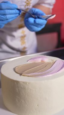 Pasta aşçısı beyaz pastanın üstüne kaşıkla beyaz krema sürüyor. Kapatın. Arkadaki masada çikolatalı pasta. Dikey video