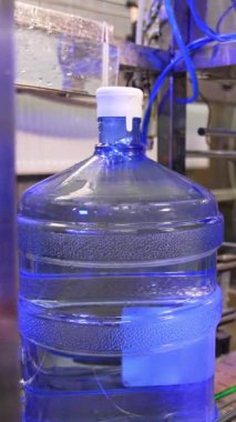 Büyük temiz su şişeleri taşıyıcı bandın yanında hareket ediyor. Makine bir şişe kaldırıyor ve işçi onu plastik torbayla kaplıyor. Dikey video