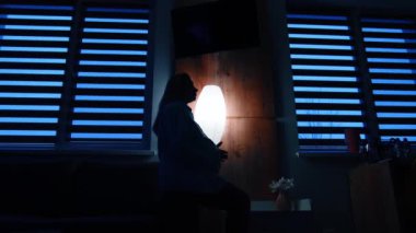 Sakin, uzun saçlı hamile bir kadın karanlık odadaki bir kanepede oturur. Gelecekteki anne karnını sevgiyle okşar. Düşük açı görünümü.