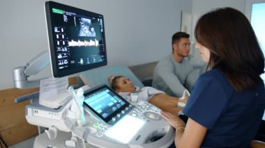 Kadın doğum uzmanı sonografi için modern ekipman üzerinde çalışıyor. Koyu renk saçlı doktor, beyaz bir kadının hamileliğini kontrol ediyor. Arkaplandaki adam.