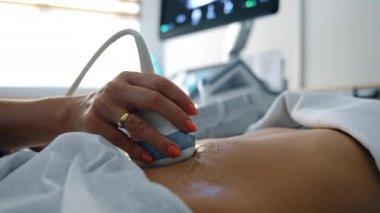 Doktor, ultrason sensörünü hamile bir hastanın jöleli karnıyla hareket ettiriyor. Kapatın. Görüntü arkaplanındaki makinenin monitörü.