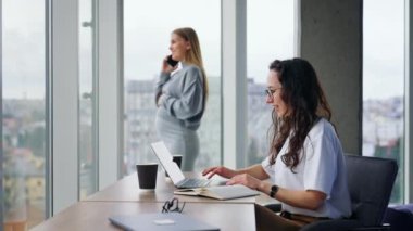 Modern geniş ofiste çalışan orta yaşlı genç bayanlar. Esmer, dizüstü bilgisayarında çalışıyor. Hamile meslektaşım pencerenin yanında telefonla konuşuyor..