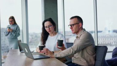 Ofis çalışanları öğle arasında çalışmaya devam ediyor. İnsanlar kahve içer ve laptopta sorunları tartışır. Arka plandaki kadın telefonunu kullanıyor..